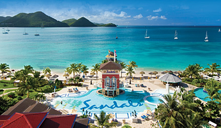 The quintessential Sandals pool and building with bright blue ocean water and rocky moutains in the background