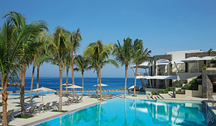 The pool area with lounge chairs and the ocean, beach and palm trees in the background at a Secrets Resort
