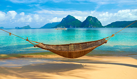 A woven hammock at the edge of crystal clear blue water with lush mountain peaks in the background